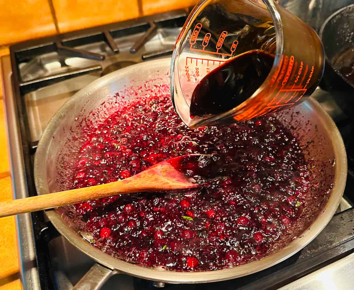 Balsamic vinegar being poured from glass measuring cup into blueberry sauce simmering in a steel skillet with a wooden spoon.