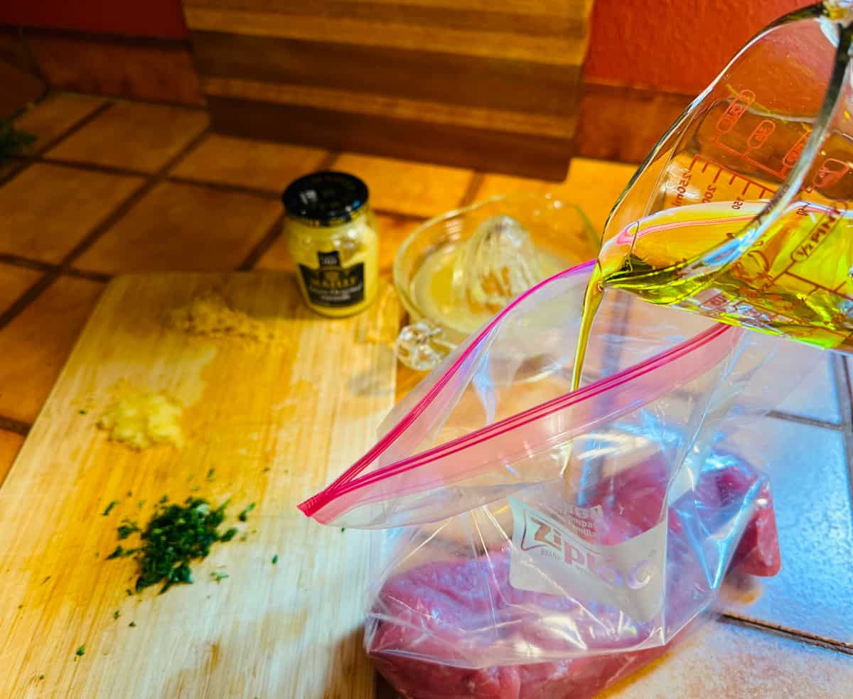 Olive oil being poured into a gallon sized ziplock bag containing raw pork tenderloin.