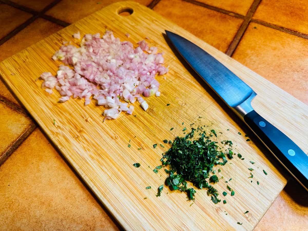 Chopped shallot and chopped thyme with a chef's knife on a wooden cutting board.