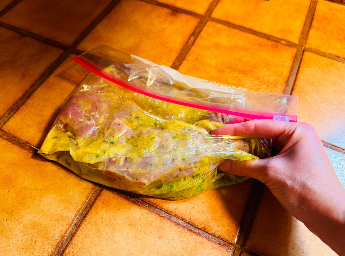 Raw pork tenderloin coated in yellow liquid flecked with thyme marinating in a gallon sized ziplock bag.