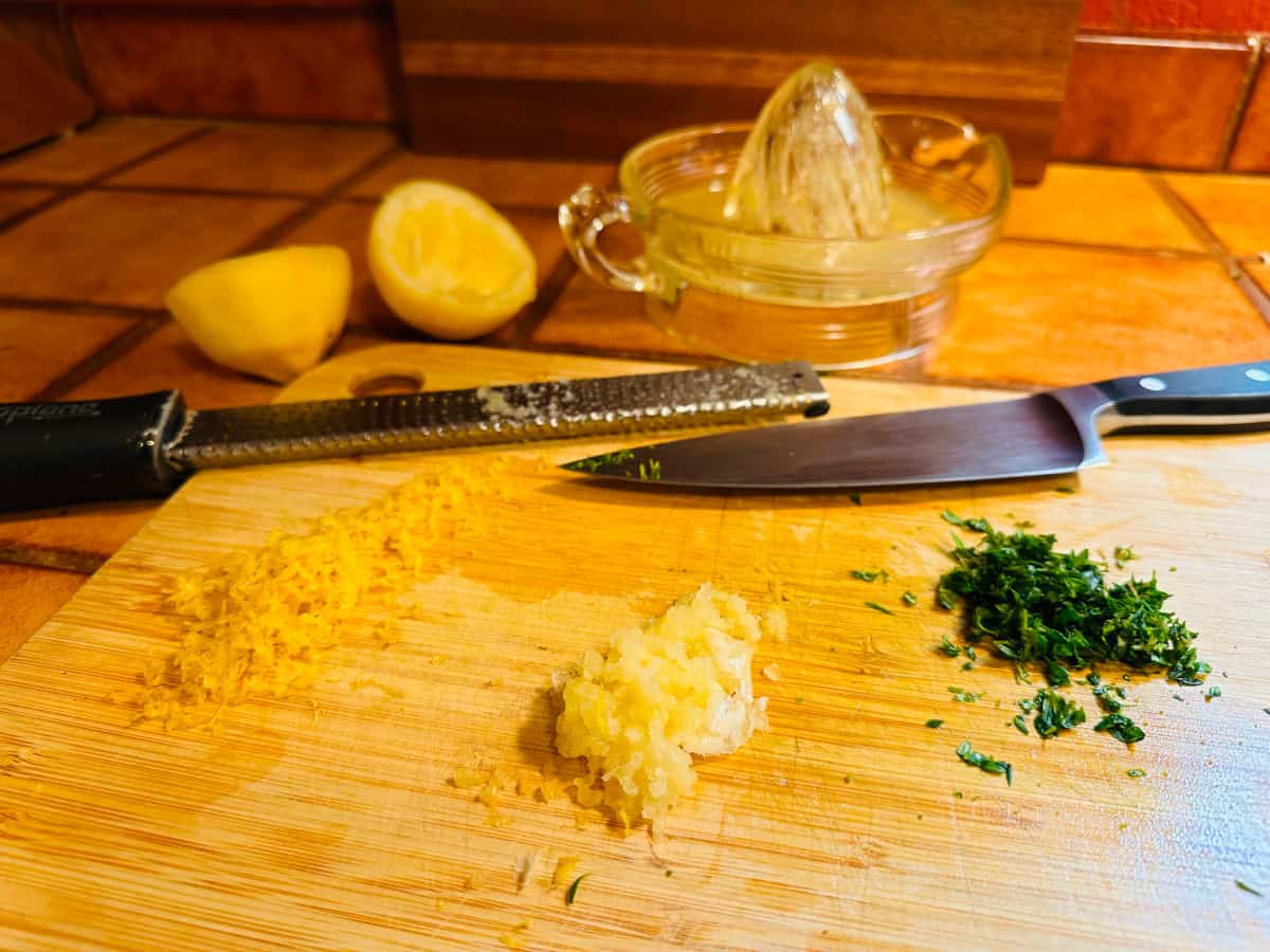 Lemon zest, grated garlic, and chopped thyme on a cutting board with a zester and a chef's knife in front of two lemon halves and a glass juicer.