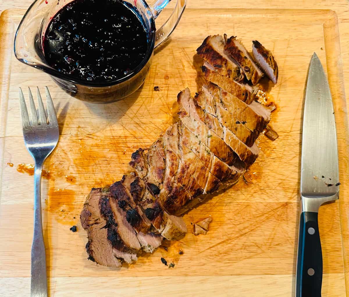 Blueberry sauce in a glass pitcher and sliced pork tenderloin between a fork and chef's knife on a wooden cutting board.