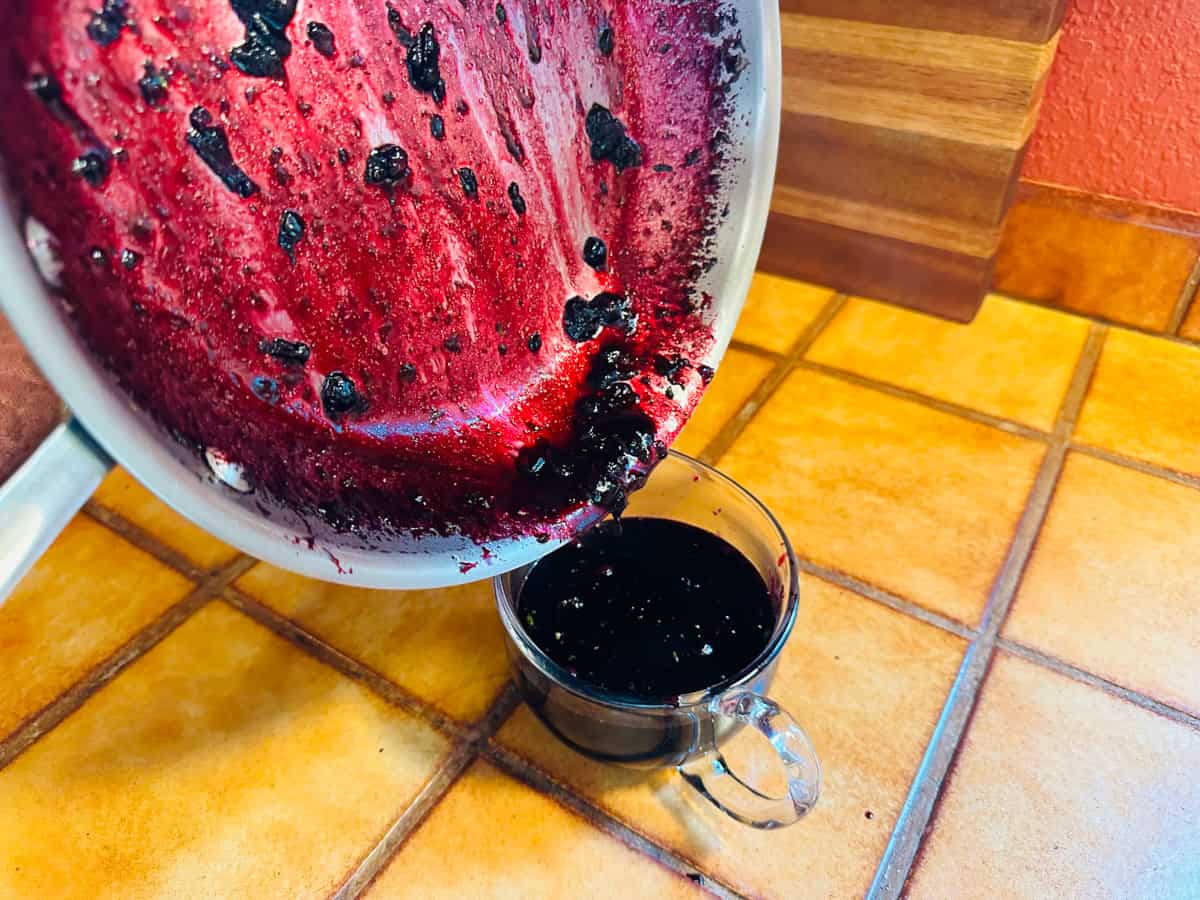 Blueberry sauce being poured from a steel skillet into a small glass pitcher.