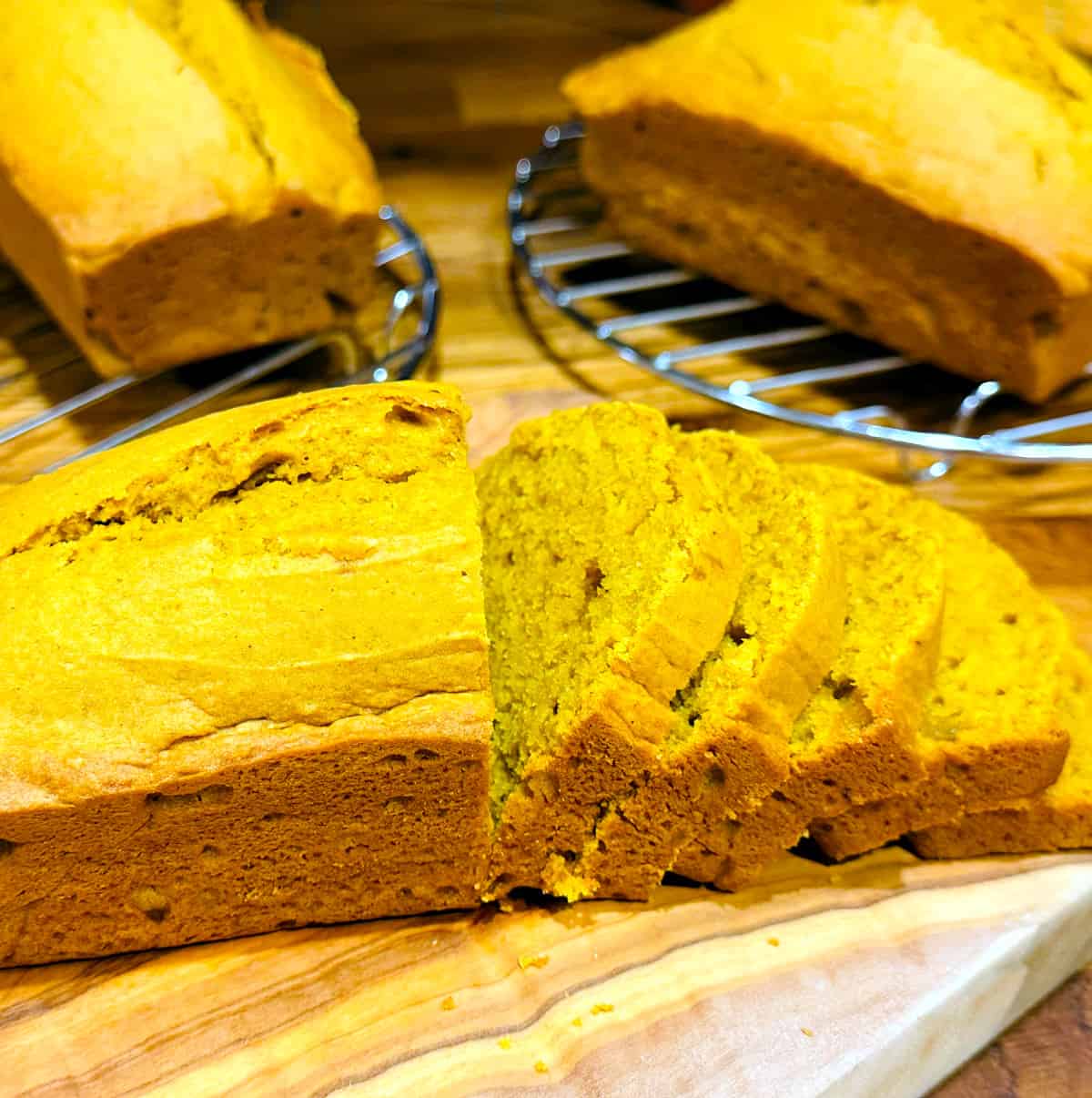 Partially sliced loaf of pumpkin bread on a wooden cutting board in front of two loaves each cooling on round wire racks.