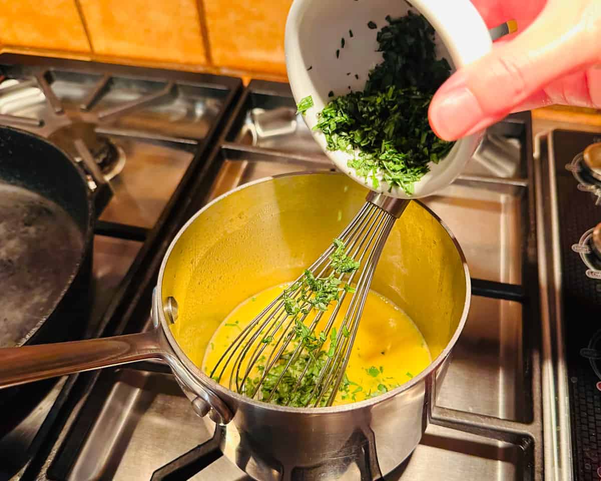 Chopped tarragon being tipped out from a small white bowl into a small steel saucepan containing yellow liquid and a whisk.