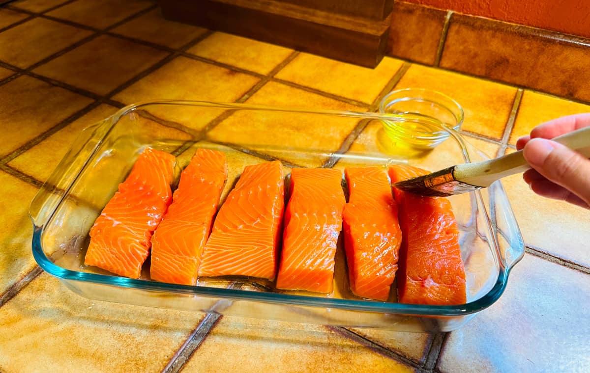 Six bright orange salmon fillets in a shallow glass casserole dish being brushed with olive oil.