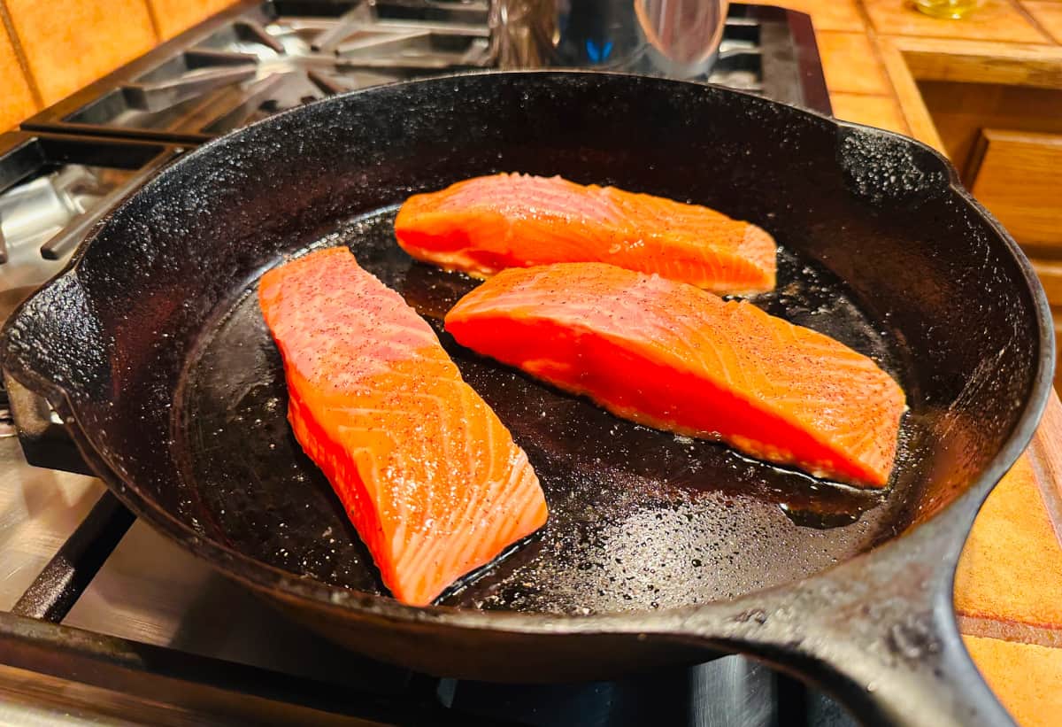 Three bright orange salmon fillets searing in a black cast iron skillet.