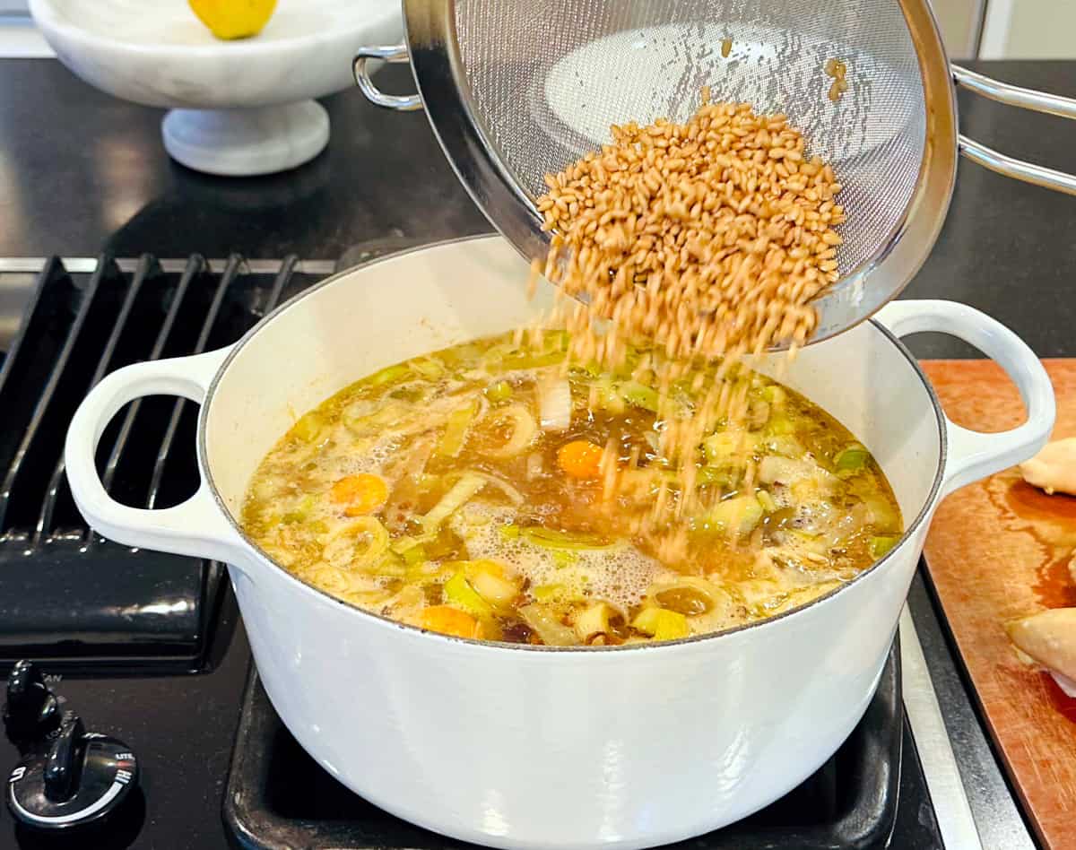 Barley being tipped from a strainer into a large white pot of simmering broth and vegetables.