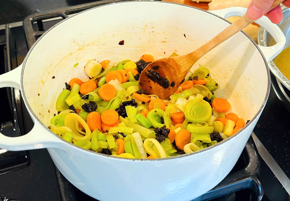 Thickly sliced leeks, carrot coins, and chopped prunes being stirred with a wooden spoon in a large white pot.