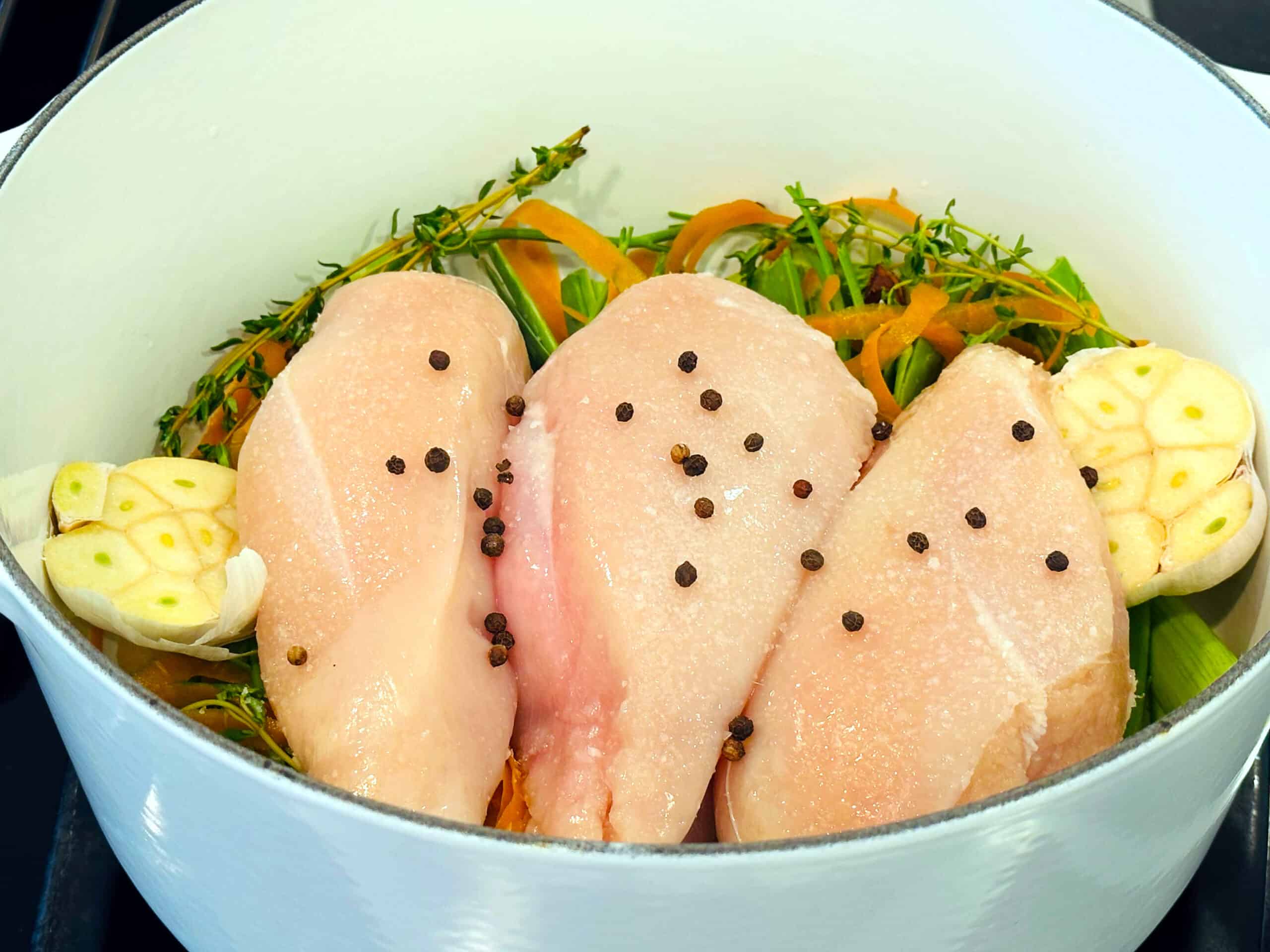 Raw chicken breasts sprinkled with whole peppercorns and salt on top of vegetables, herbs, and garlic in a large white pot.