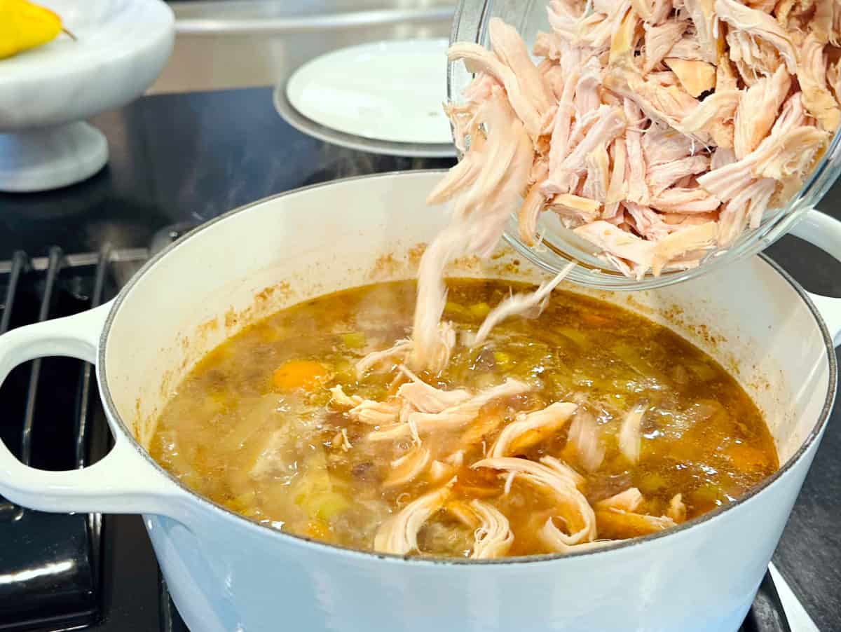 Shredded chicken being tipped from a glass bowl into cock-a-leekie soup in a large white pot.