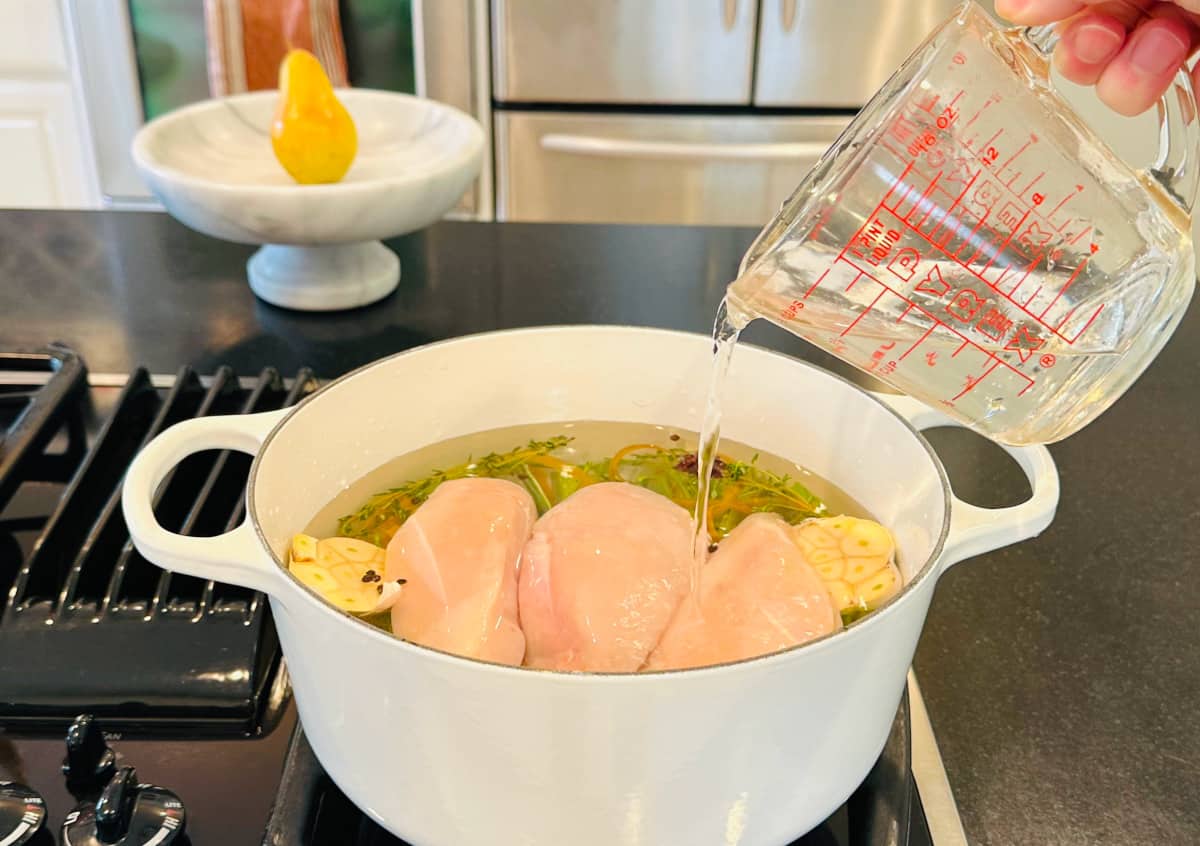 Water being poured from a glass measuring cup over raw chicken breasts, vegetables, and herbs in a large white pot.