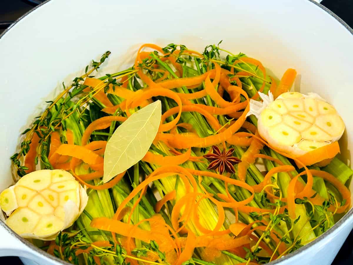 Halved head of garlic, carrot peels, thyme sprigs, leek tops, dried bay leaf, and star anise in a large white pot.