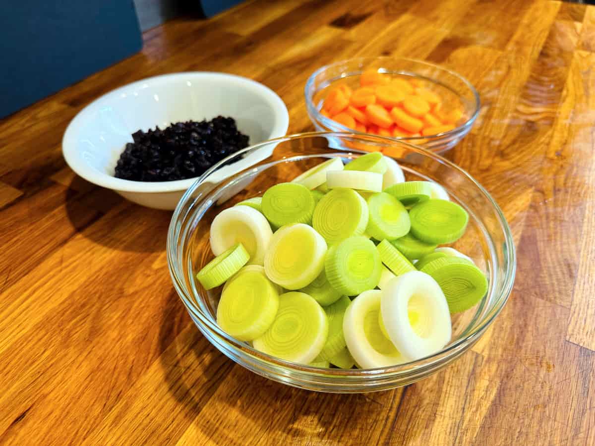 Thick slices of leek in a medium glass bowl, carrot coins in a small glass bowl, and chopped prunes in a small white ceramic bowl.