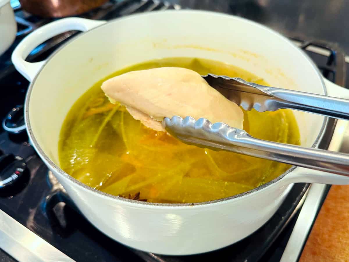 Cooked chicken breast being removed with metal tongs from broth in a large white pot.