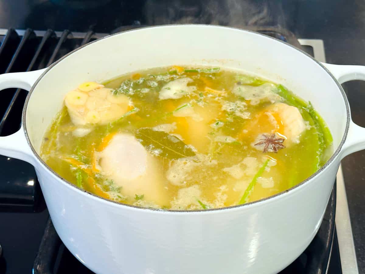 Chicken breasts, vegetables, and herbs in pale yellow liquid simmering in a large white pot.