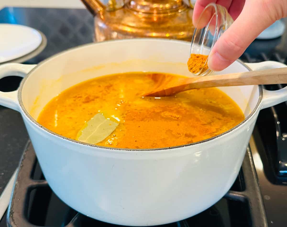 Curry powder being tipped from a small glass bowl into orange liquid with a bay leaf and a wooden spoon in a white pot on the stove.