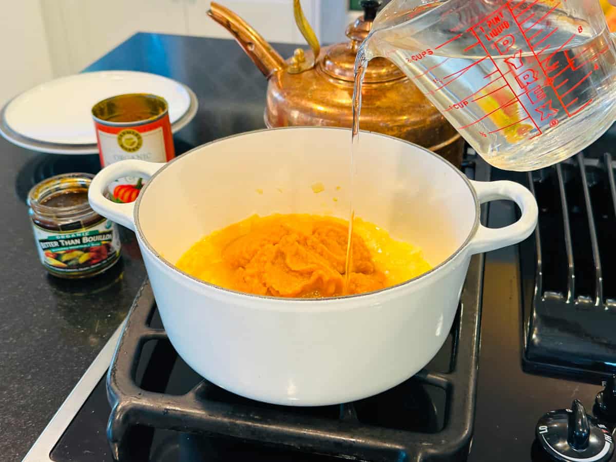 Water being poured into pumpkin puree and cooked onions in a white pot on the stove.