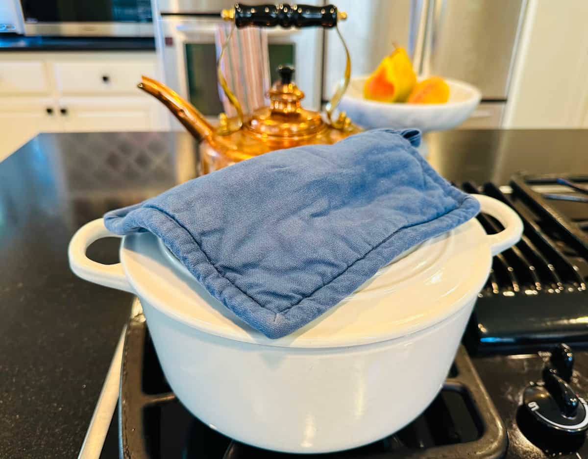 A white pot sitting on the stove with a blue pot holder on top of the lid.
