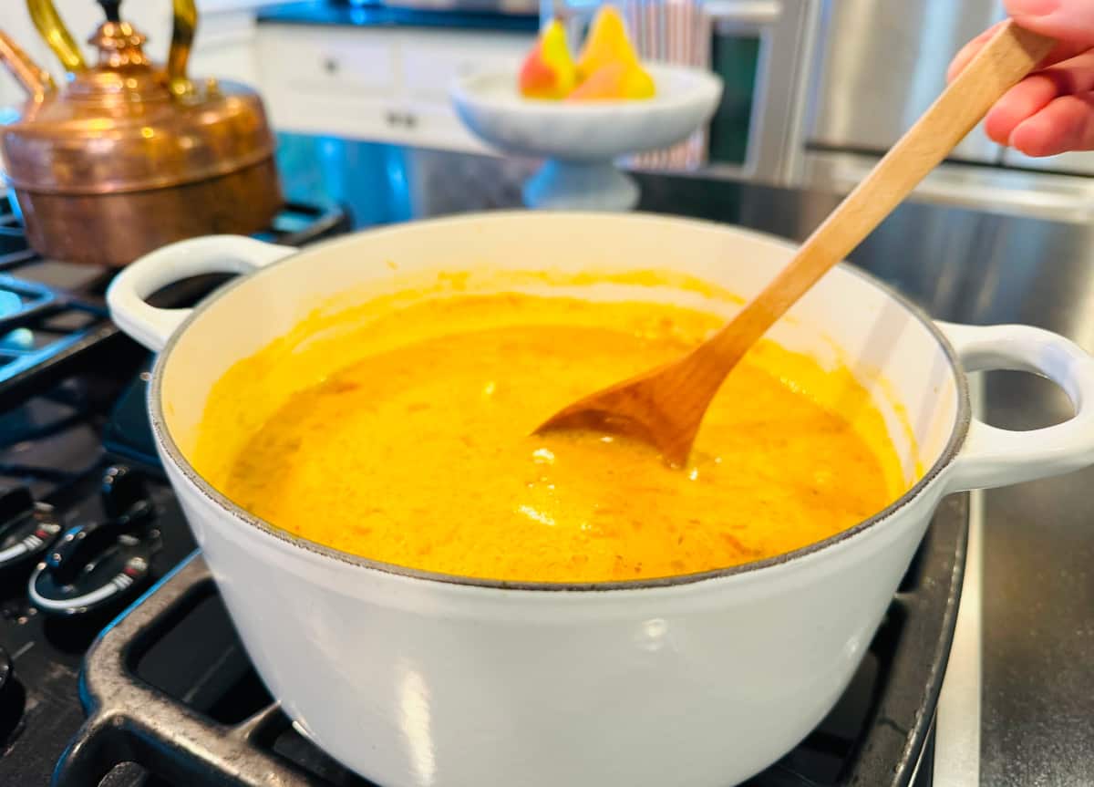 Curried pumpkin soup being stirred with a wooden spoon in a white pot on the stove.