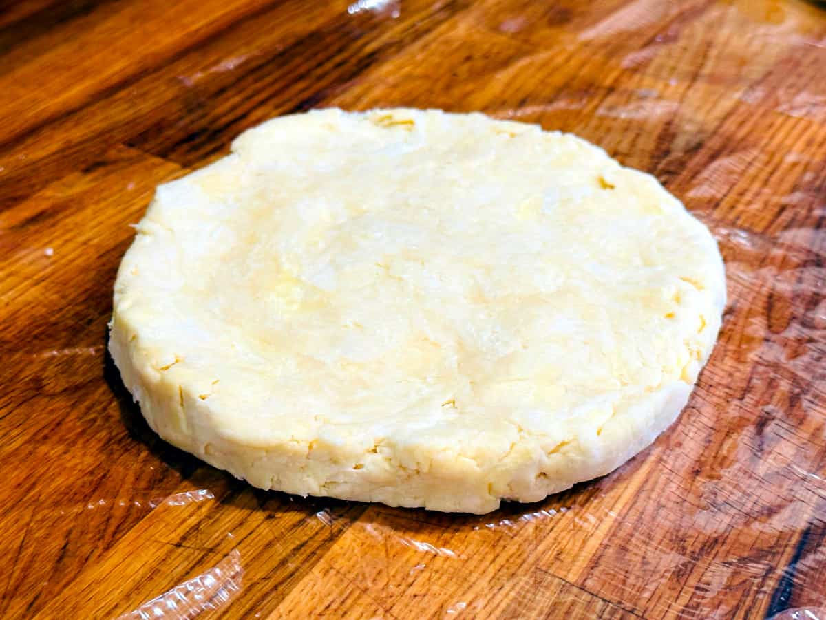 Disk of butter crust dough on plastic wrap sitting on a butcher block counter.