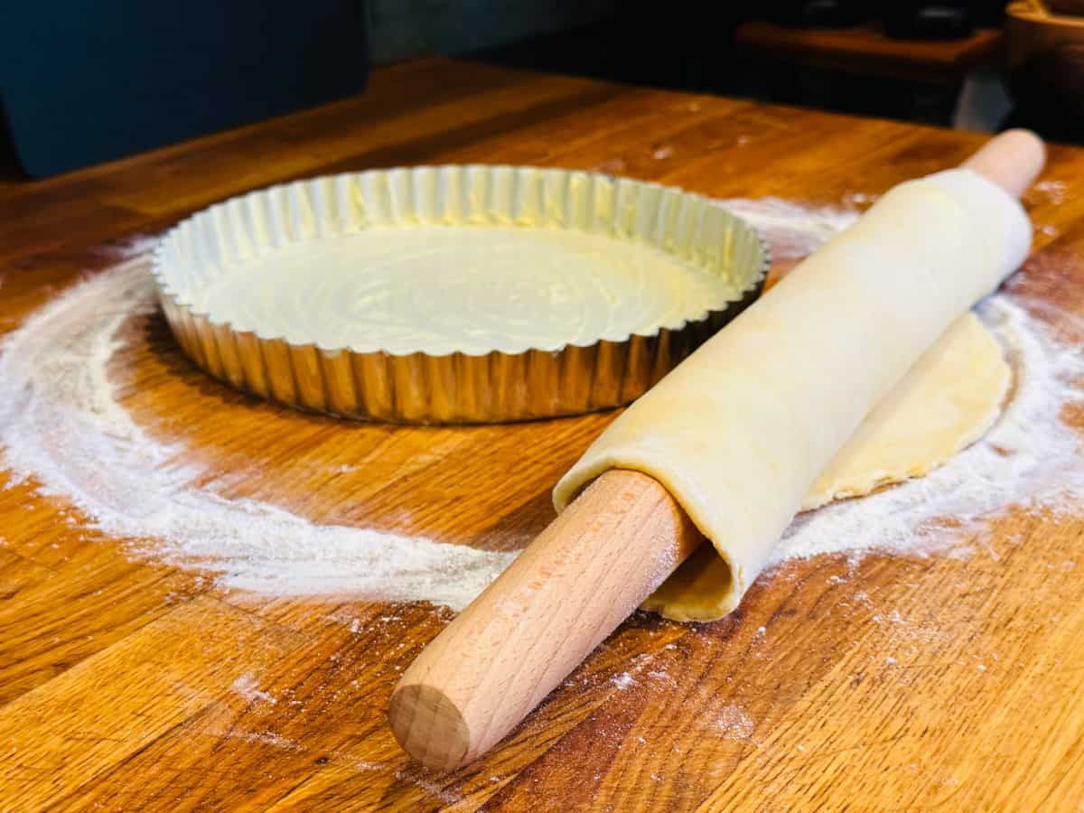 Rolled out crust dough gathered onto a rolling pin next to a greased tart pan.