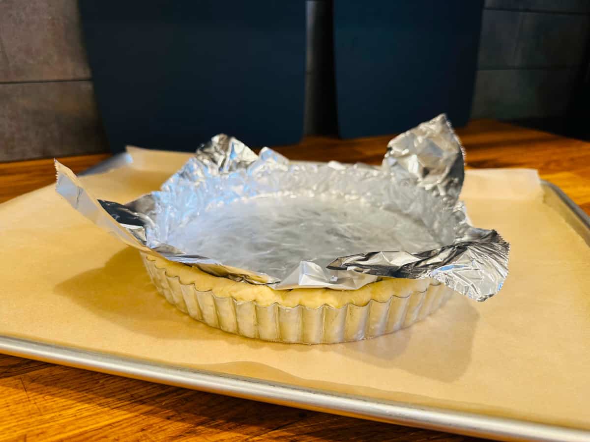 Aluminum foil lining unbaked crust dough in a tart pan sitting on a parchment covered baking sheet.