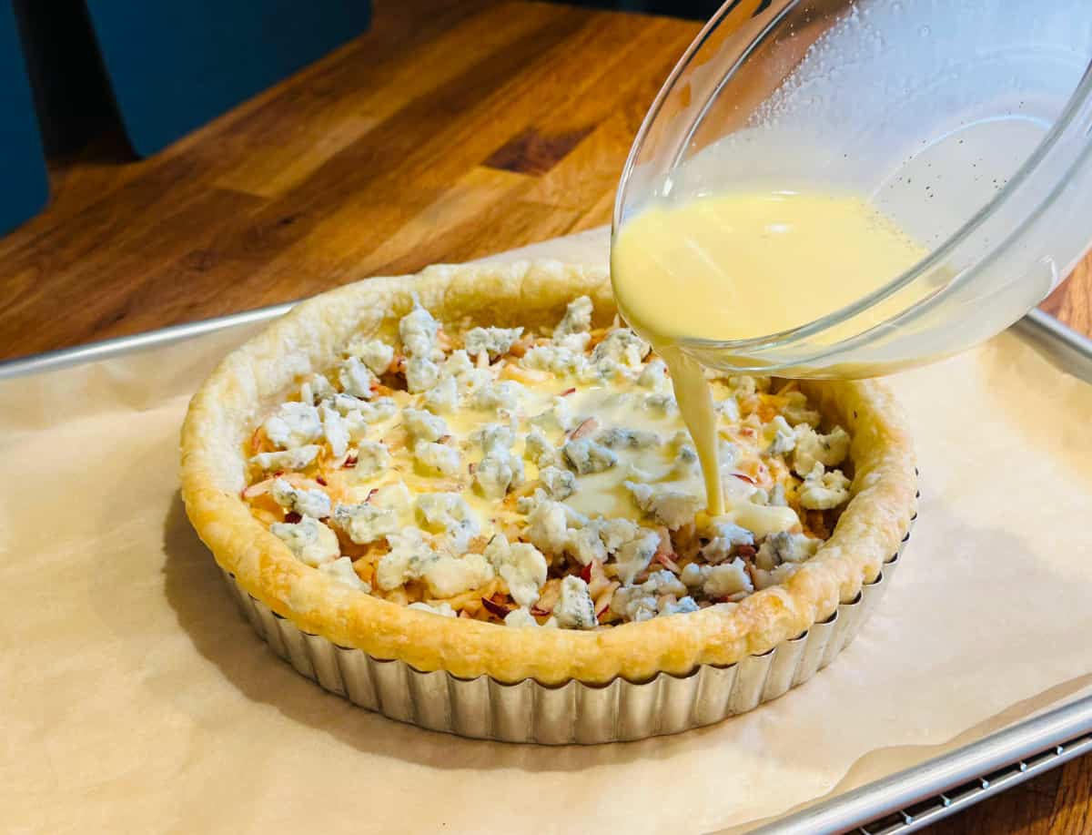 Beaten eggs and cream being poured over small pieces of Gorgonzola dolce and shredded apple in a partially baked quiche shell.