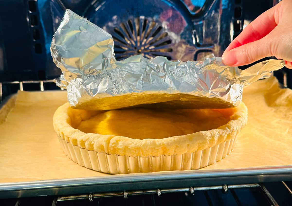 Aluminum foil being removed from partially baked crust in a tart pan while baking in a blue oven.