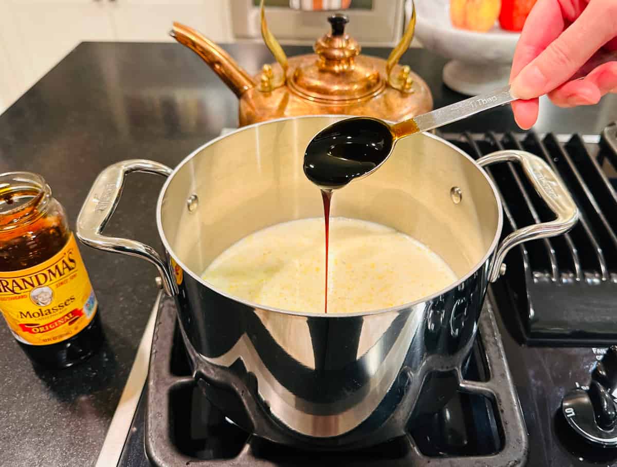 Molasses being poured from a tablespoon into milk and cornmeal in a steel saucepan on the stove.