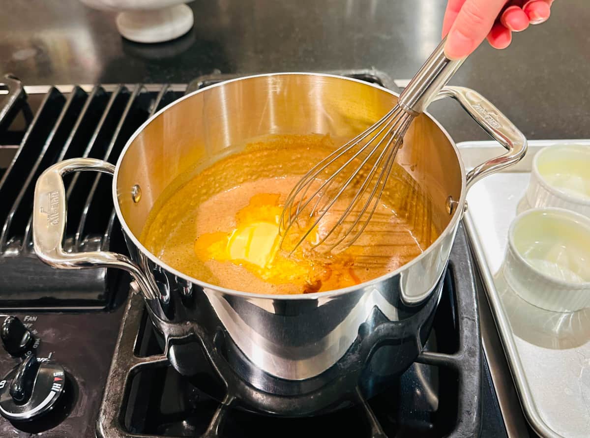 Melting butter and vanilla extract being whisked into light brown mixture in a steel saucepan.