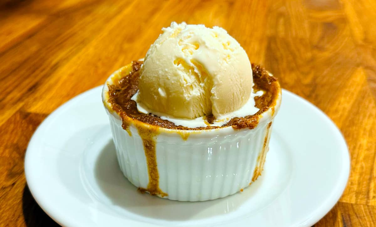 Hasty pudding topped with a scoop of vanilla ice cream in a white porcelain ramekin on a small white plate.
