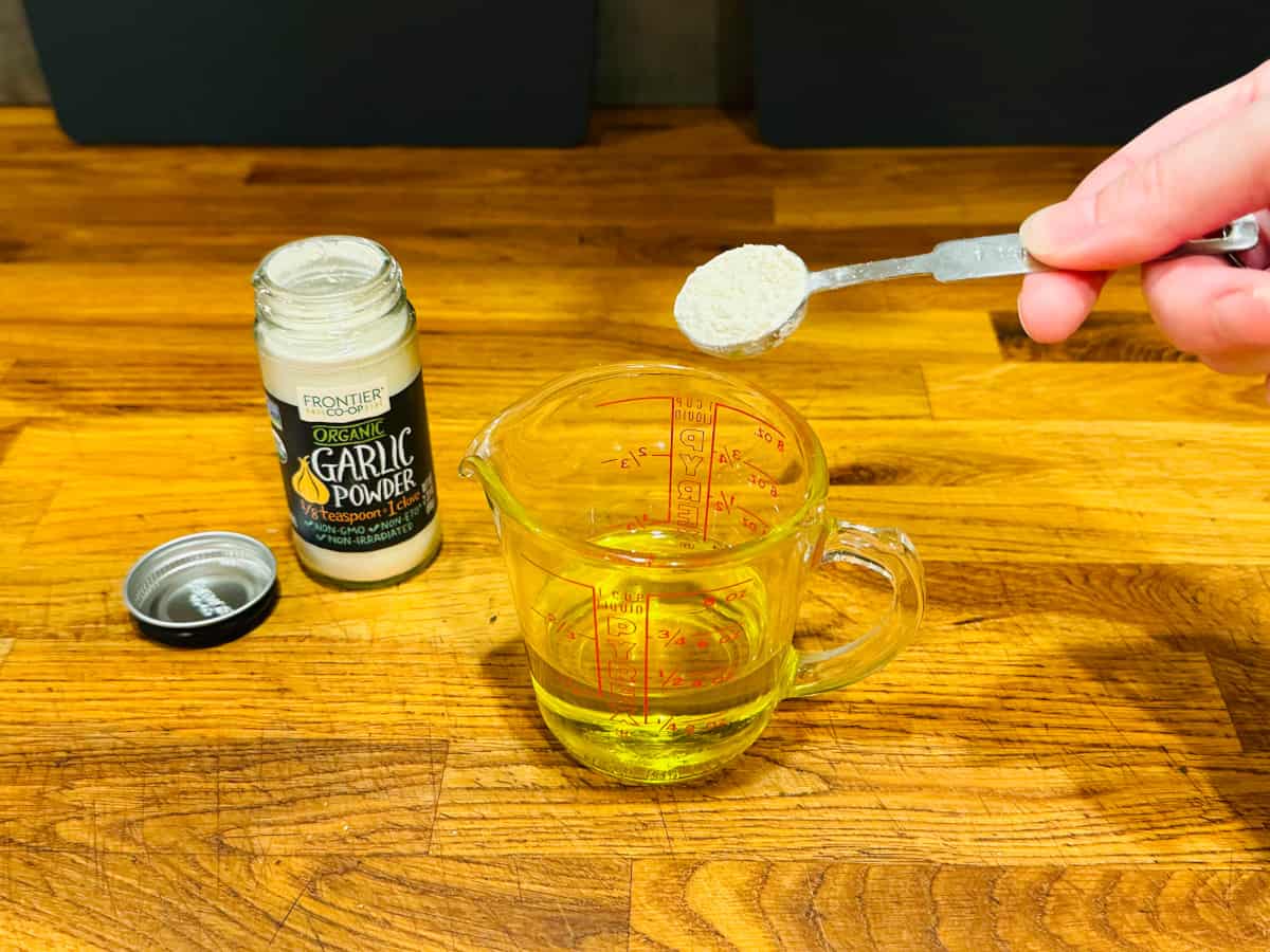 White powder in a metal teaspoon being held over a measuring cup containing olive oil next to a jar of garlic powder.