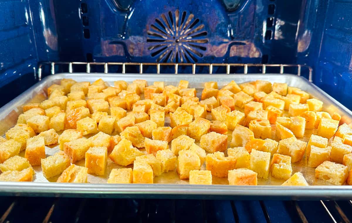Homemade croutons roasting on a metal baking sheet in a blue oven.