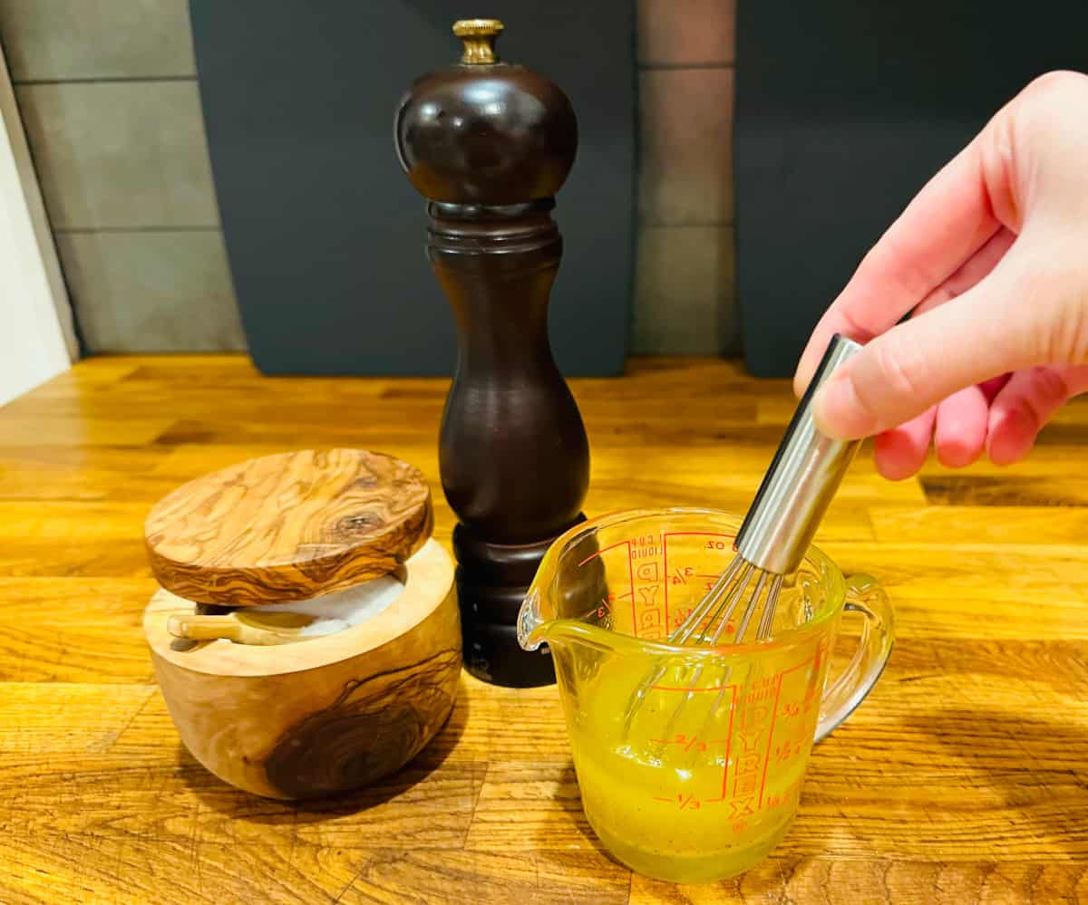 Olive oil and seasonings being stirred with a small metal whisk in a glass measuring cup next to a salt dish and pepper mill.