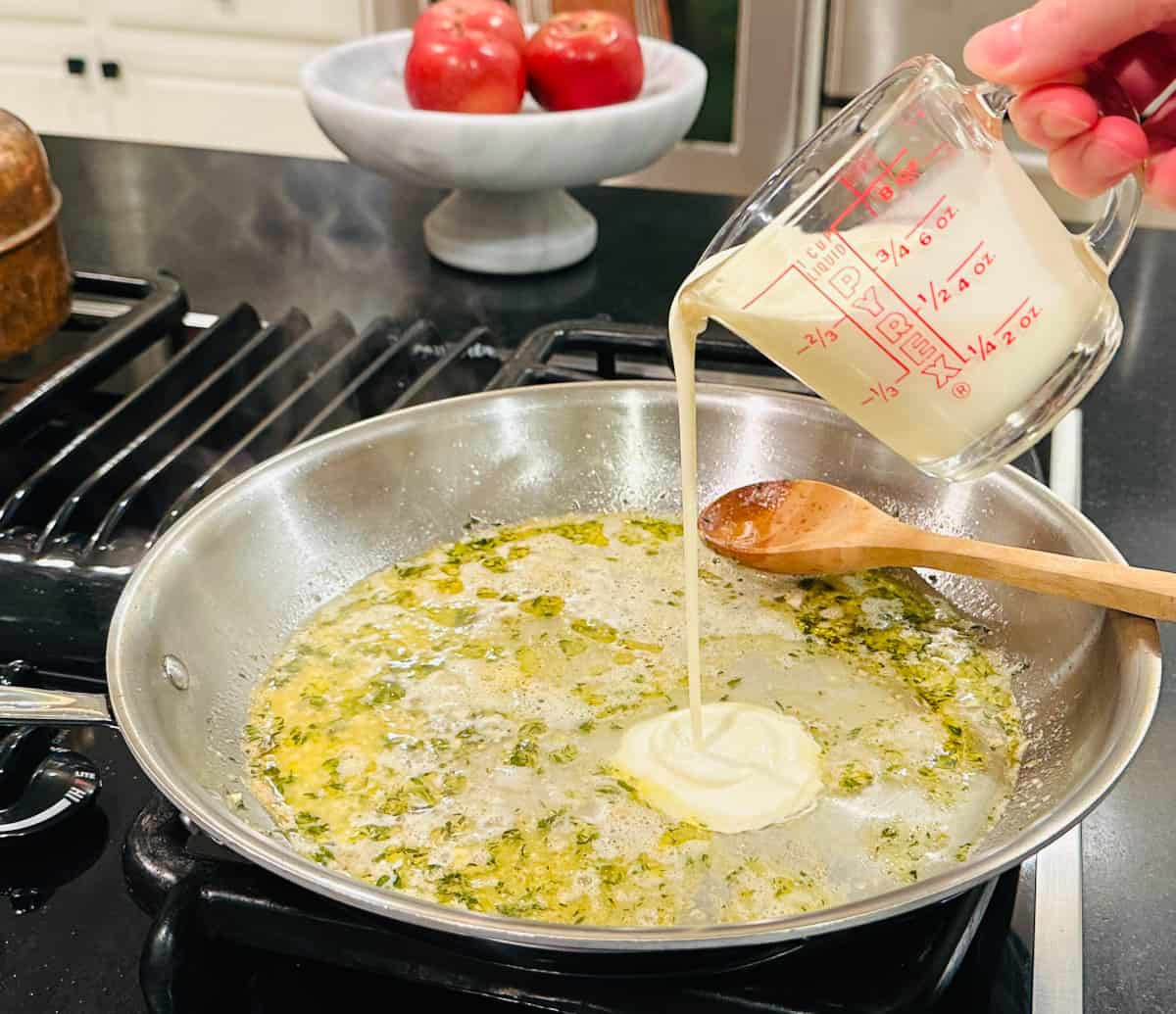 Cream being poured from a glass measuring cup into a large steel skillet containing white wine, melted butter, thyme, and garlic along with a wooden spoon.