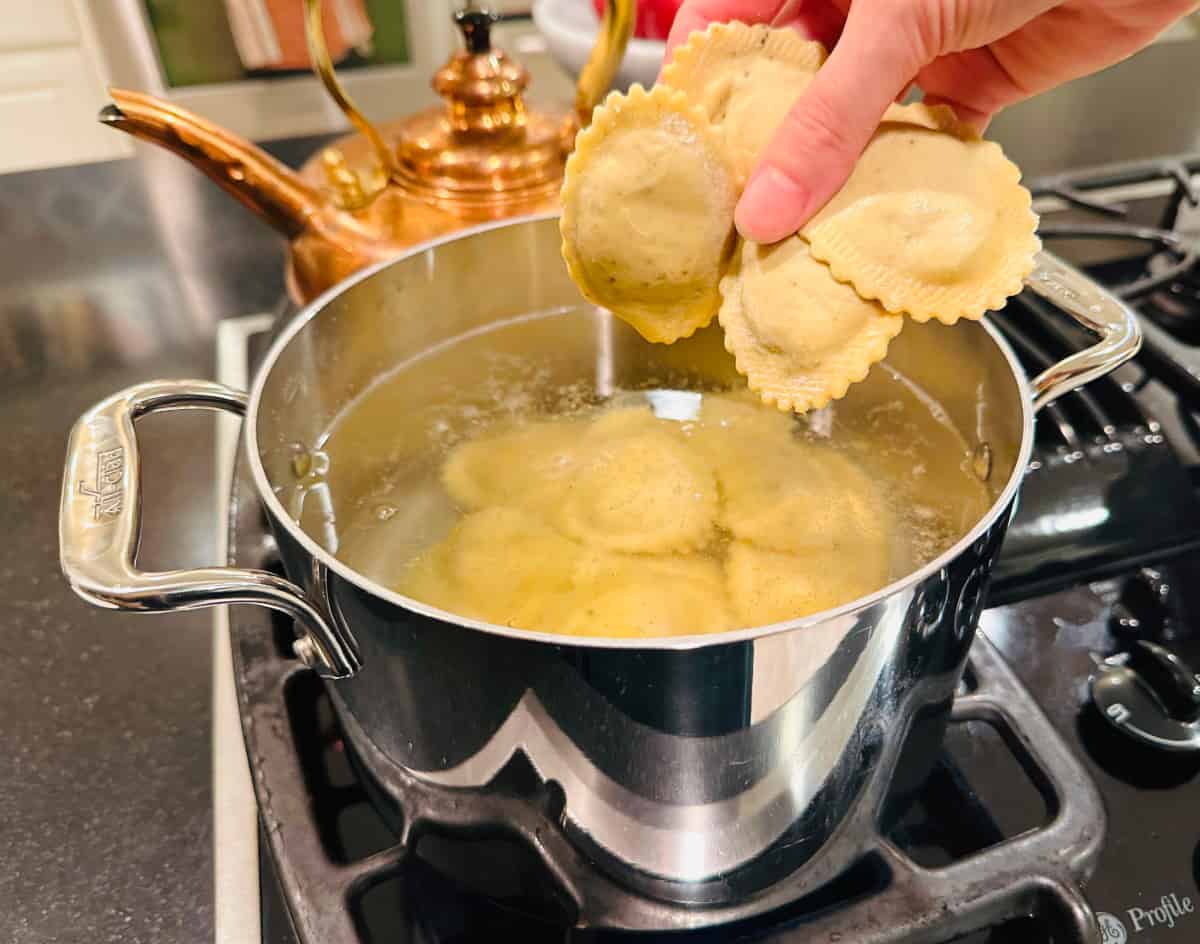 A handful of ravioli being held over a medium steel pot of simmering water and ravioli.