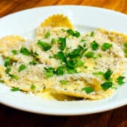 Mushroom ravioli with cream sauce sprinkled with shredded parmesan and chopped fresh parsley in a shallow white bowl.