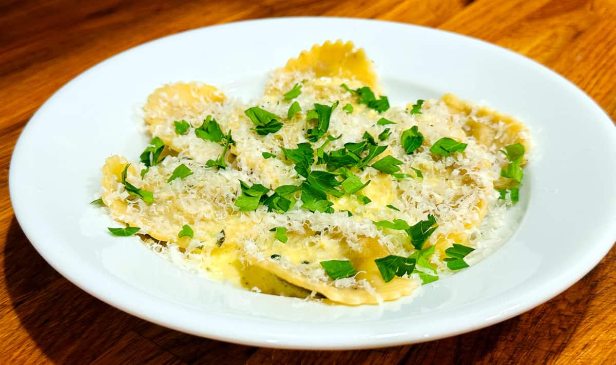 Mushroom ravioli with cream sauce sprinkled with shredded parmesan and chopped fresh parsley in a shallow white bowl.