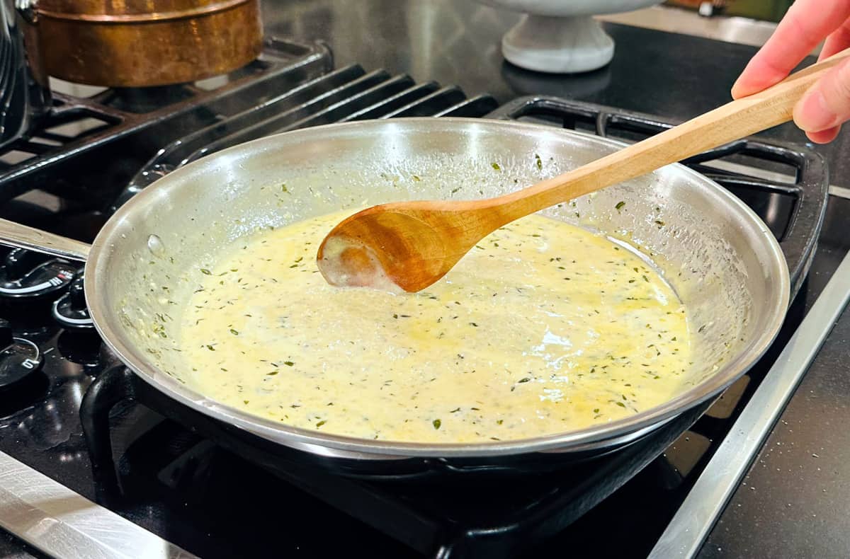 Cream sauce flecked with thyme being stirred with a wooden spoon in a large steel skillet.