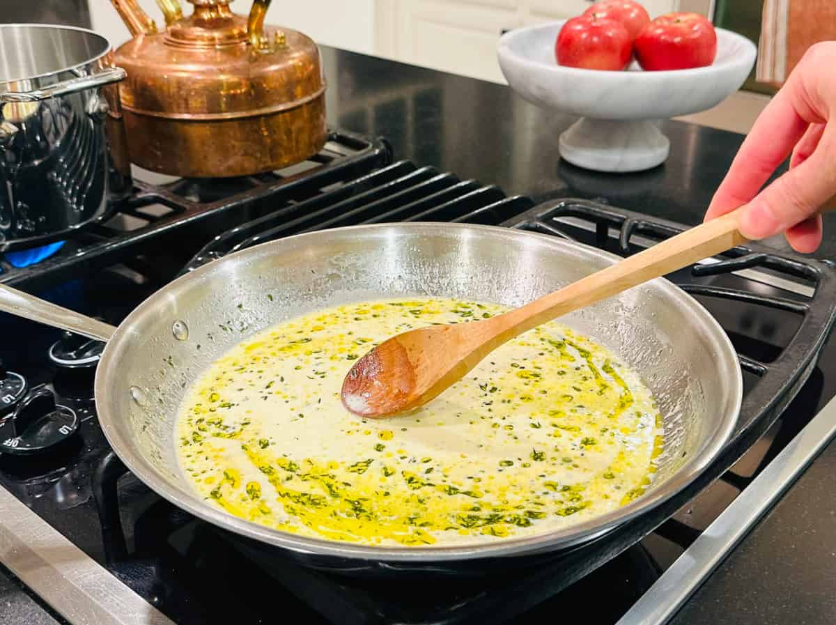Cream sauce flecked with thyme being stirred with a wooden spoon in a large steel skillet.