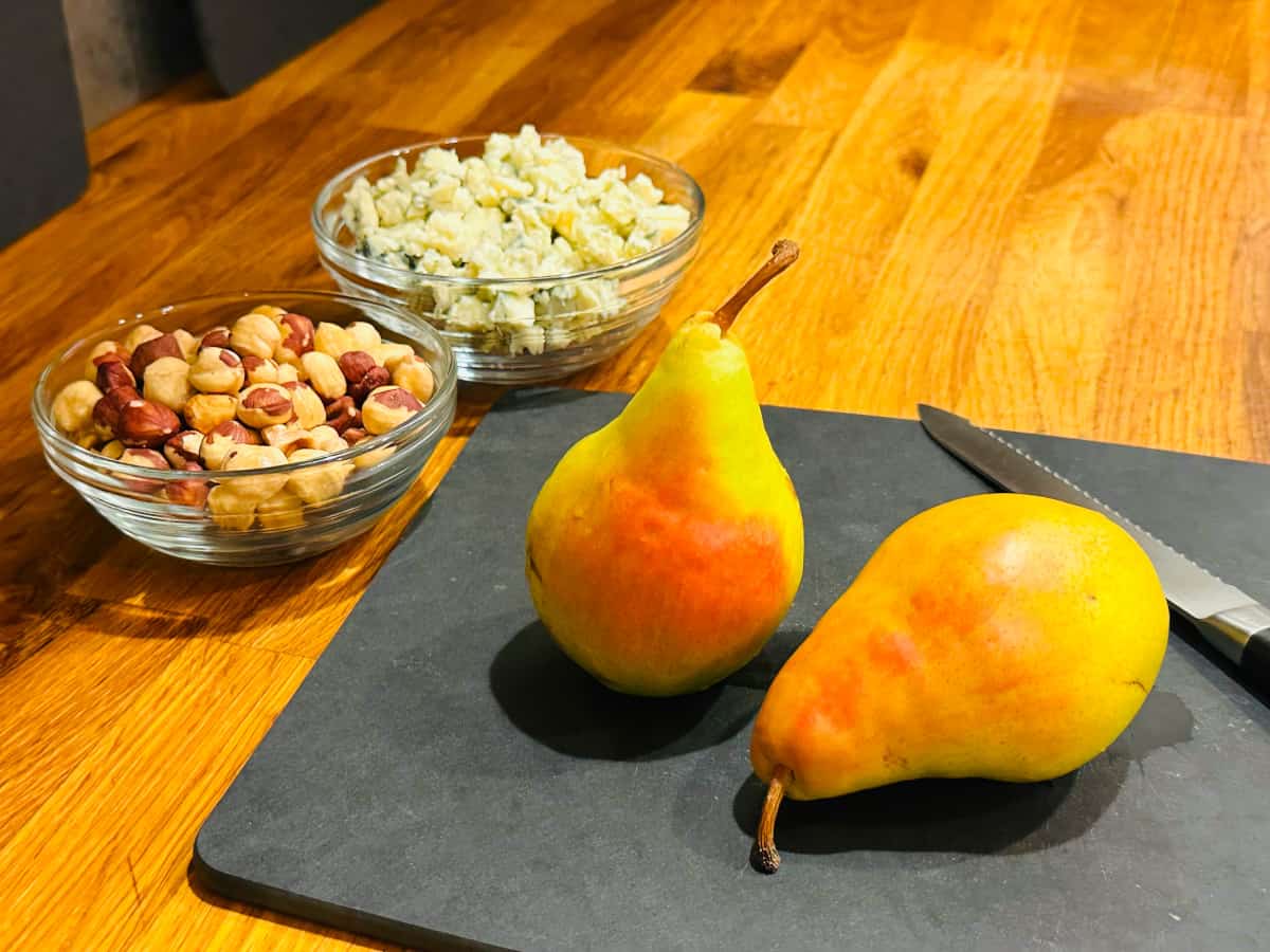 Hazelnuts in a small glass bowl, crumbled blue cheese in a small glass bowl, and two green and pink colored pears on a black cutting board with a serrated knife.