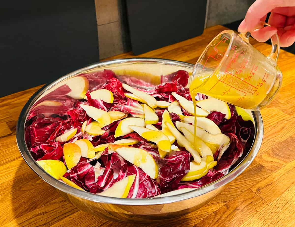 Yellow salad dressing being poured from a glass measuring cup over torn radicchio and sliced pears in a large metal bowl.