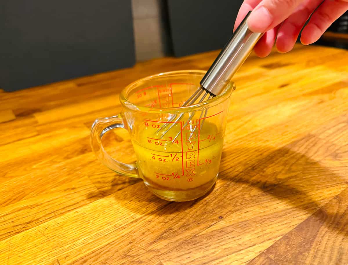 Mixing olive oil, vinegar, and mustard in a glass measuring cup with a small metal whisk.