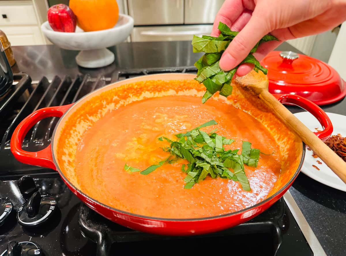 Chopped basil being sprinkled into orange colored sauce in a large red braiser on the stove.
