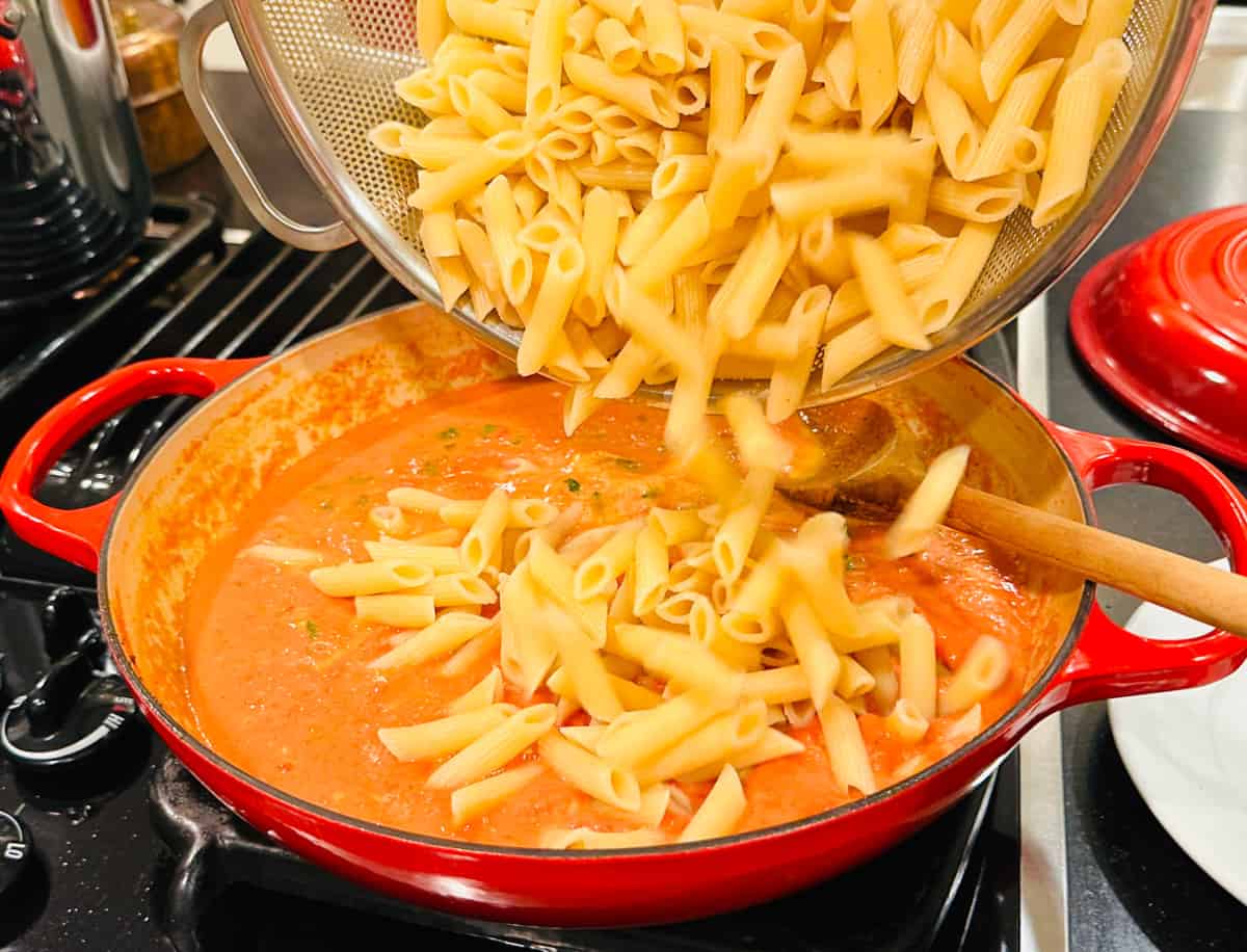 Cooked penne pasta being tipped from a colander into orange colored sauce in a large red braiser on the stove.