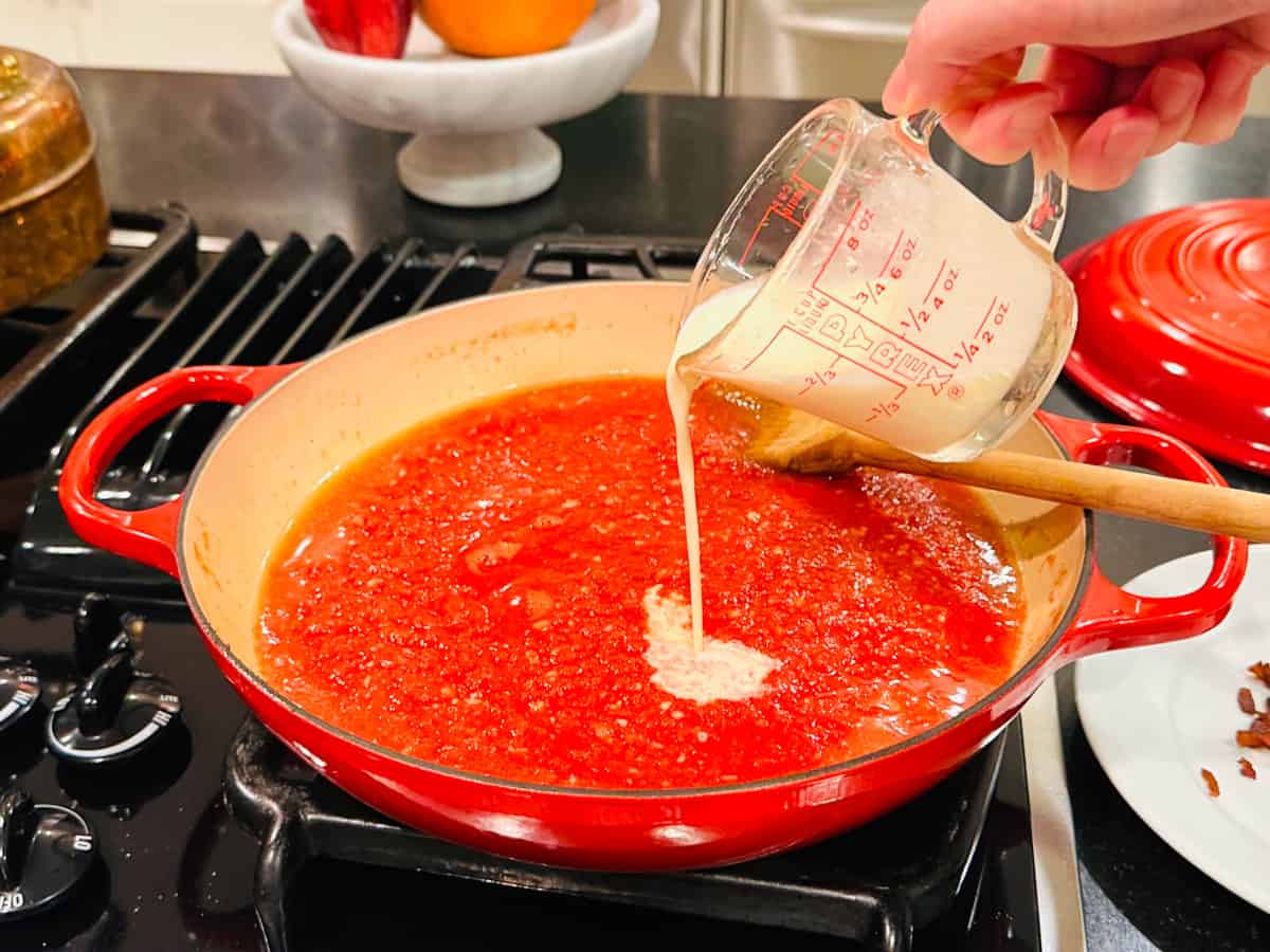 Cream being poured from a glass measuring cup into chopped tomatoes in a large red braiser on the stove.