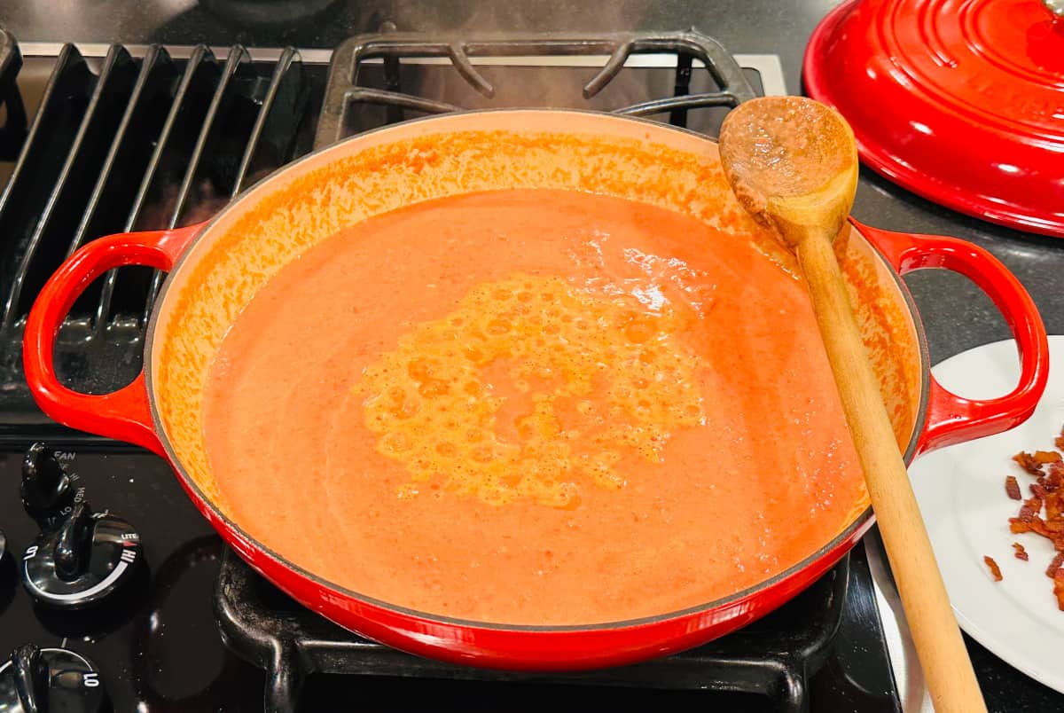 Orange colored sauce simmering in a large red braiser with a big wooden spoon resting along the edge.