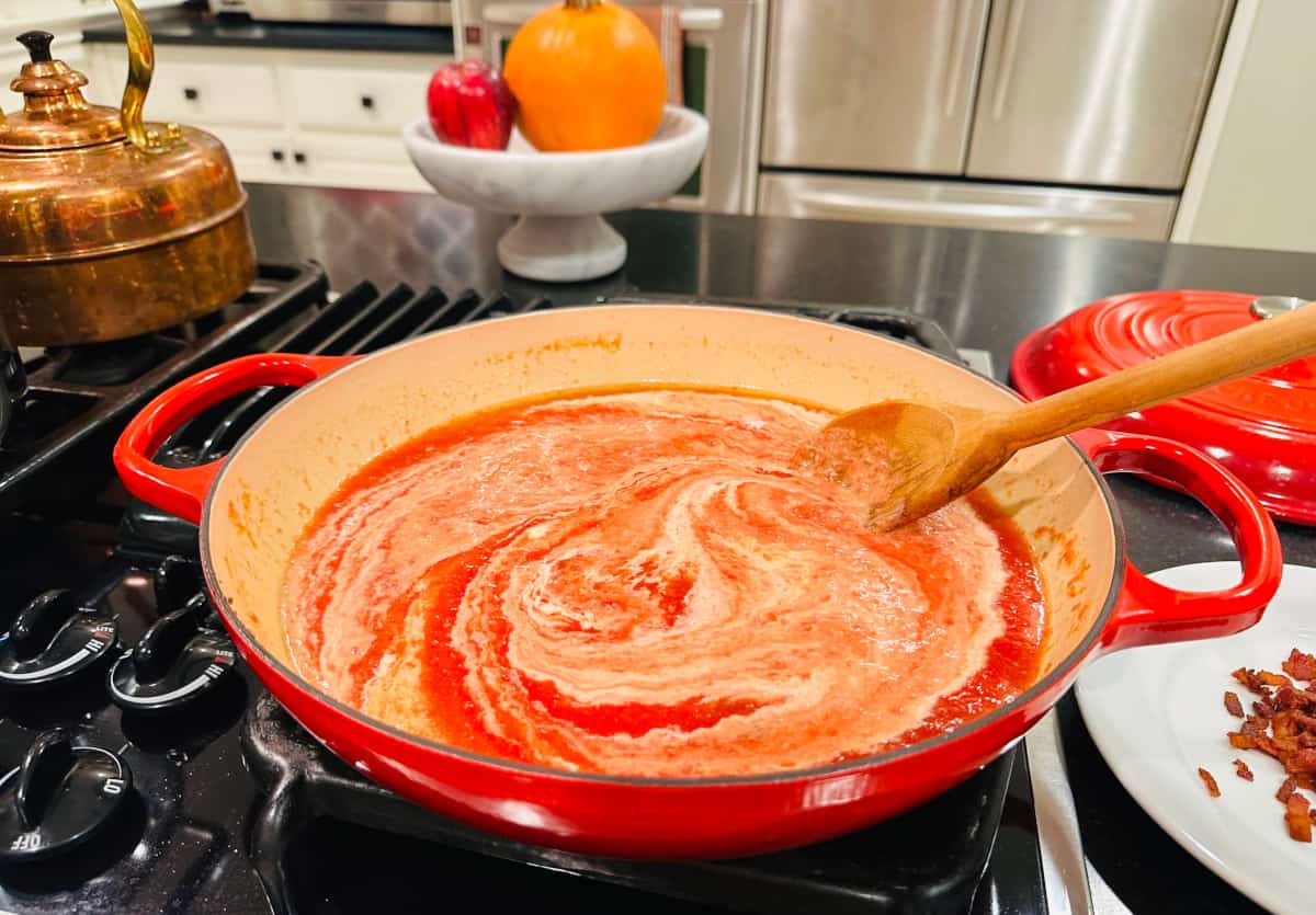 Cream being stirred into chopped tomatoes in a large red braiser on the stove.