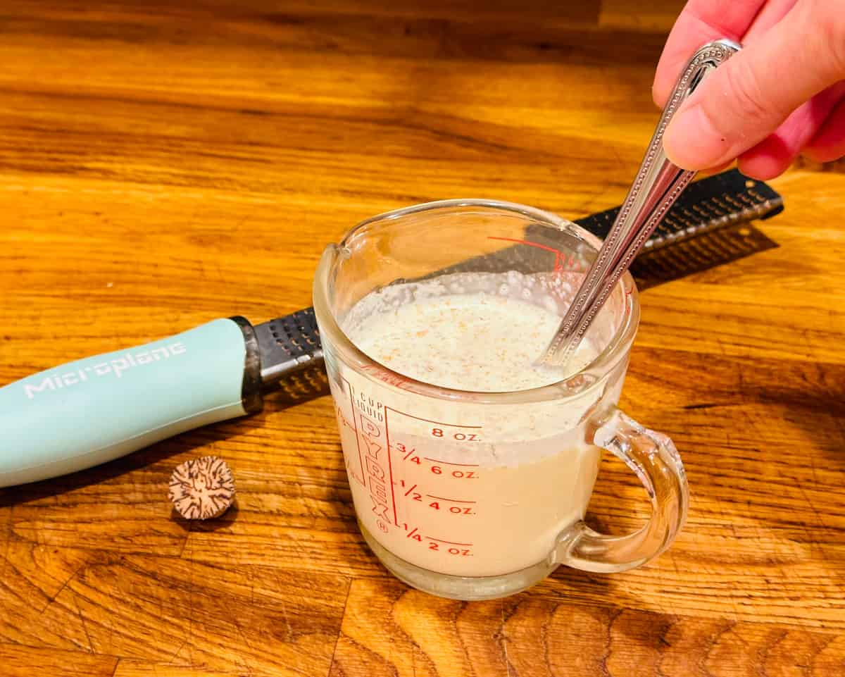Cream flecked with grated nutmeg being stirred in a glass measuring cup with a metal spoon next to a microplane grater and a piece of nutmeg.